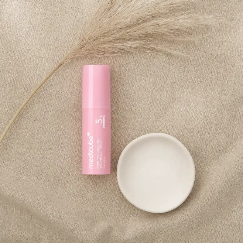 Overhead flat-lay of the medicube balm stick on oat linen beside a small white ceramic dish and a dried grass stem