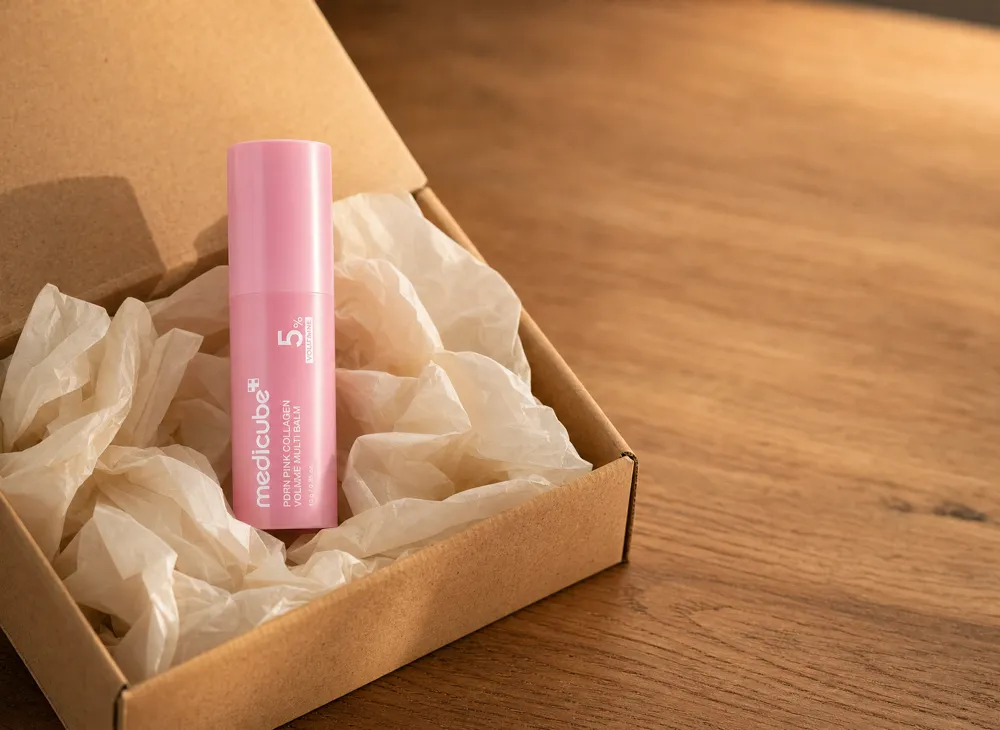 The medicube balm stick nested in cream tissue paper inside an open kraft shipping box on an oak table
