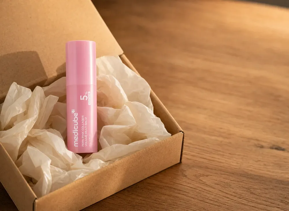 The medicube balm stick nested in cream tissue paper inside an open kraft shipping box on an oak table