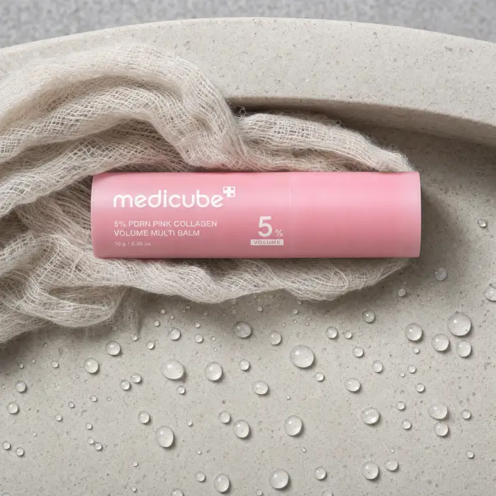 The medicube balm stick beside a damp muslin cloth and water droplets on a pale stone basin edge