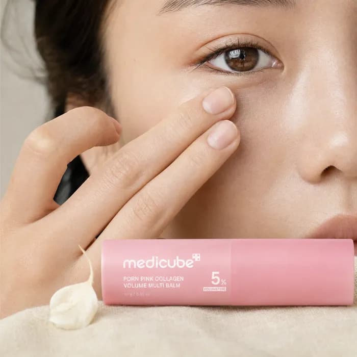 Two fingertips tapping balm into the skin under an eye, with the medicube stick resting below