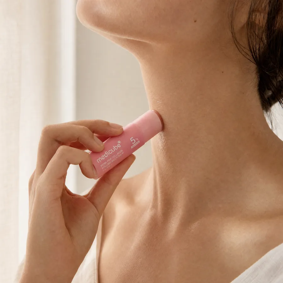 A woman holding the pink medicube balm stick against her neck in front of a pale cream curtain
